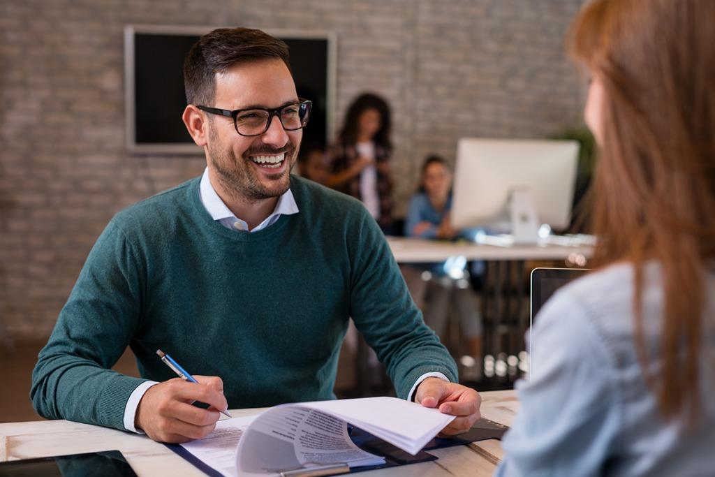 Male interviewer smiling and making notes while the female interviewee (face not visible) responds to his question