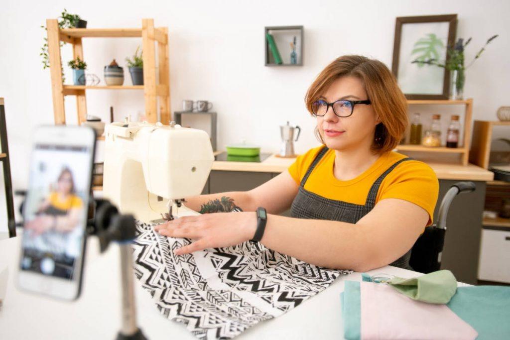 Disabled young dressmaker in eyeglasses using sewing machine while giving online class on sewing