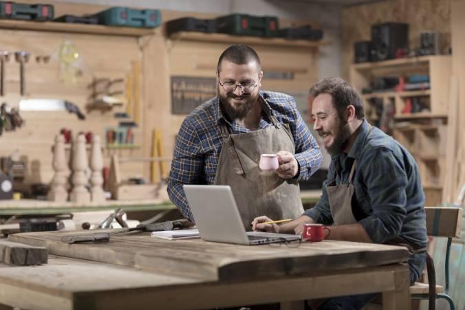 two men in a wood workshop looking at laptop smiling