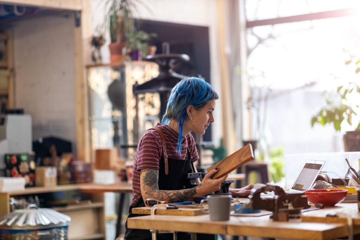 A woman in a workshop holding a block of wood and looking at a laptop