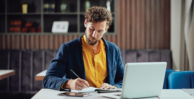 A man at a table writing on a notepad with an open laptop in front of him