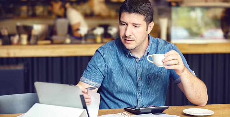 Man sipping a cup of coffee, in a coffee shop looking at a laptop screen