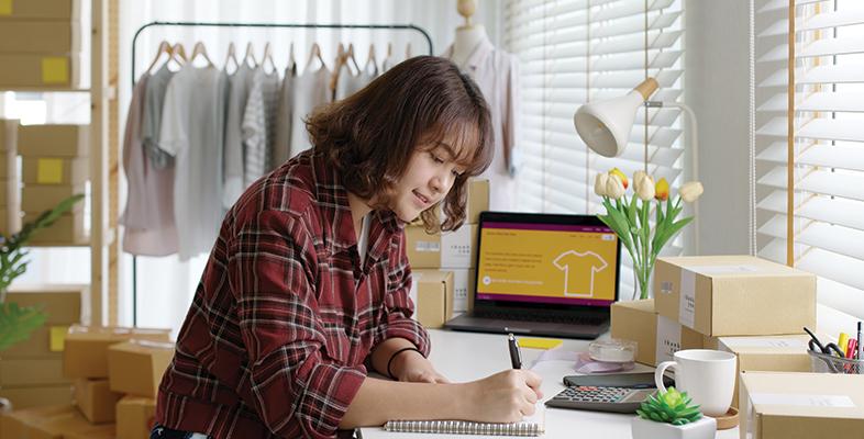A women sat working at a desk on a notepad using a pencil, laptop sat open behind her