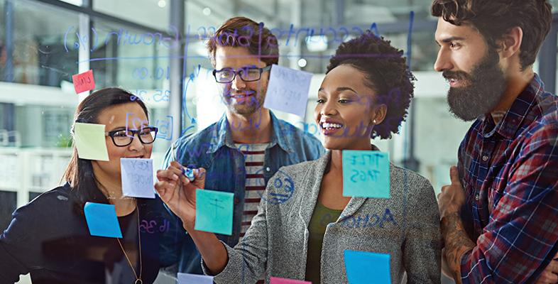 Looking through a glass window at a team of four people, two men and two women who are putting post-it notes on the other side of the glass
