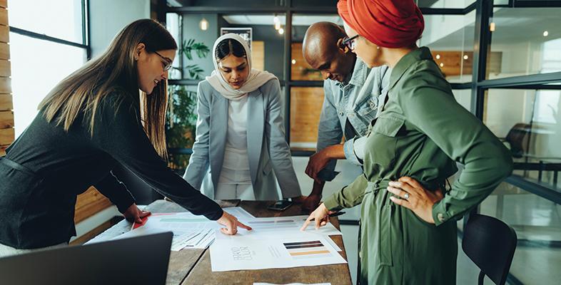 Three woman and a man standing in a modern office around a table in discussion over a document