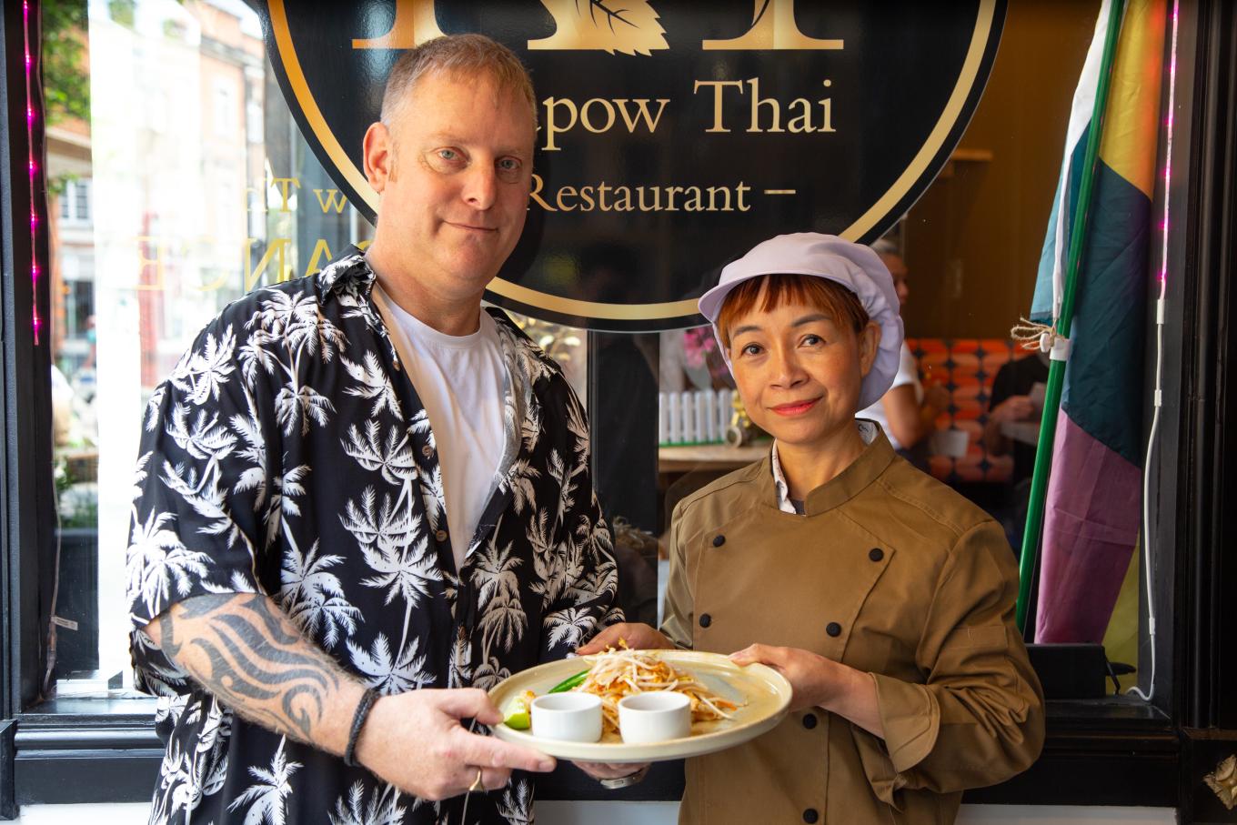Phil Williams, co-founder of Krapow Thai, an authentic Thai food restaurant. He is standing next to his partner holding a plate of food