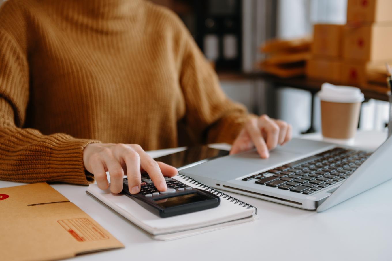 A lady sat at a desk with a laptop, using a calculator and drinking coffee