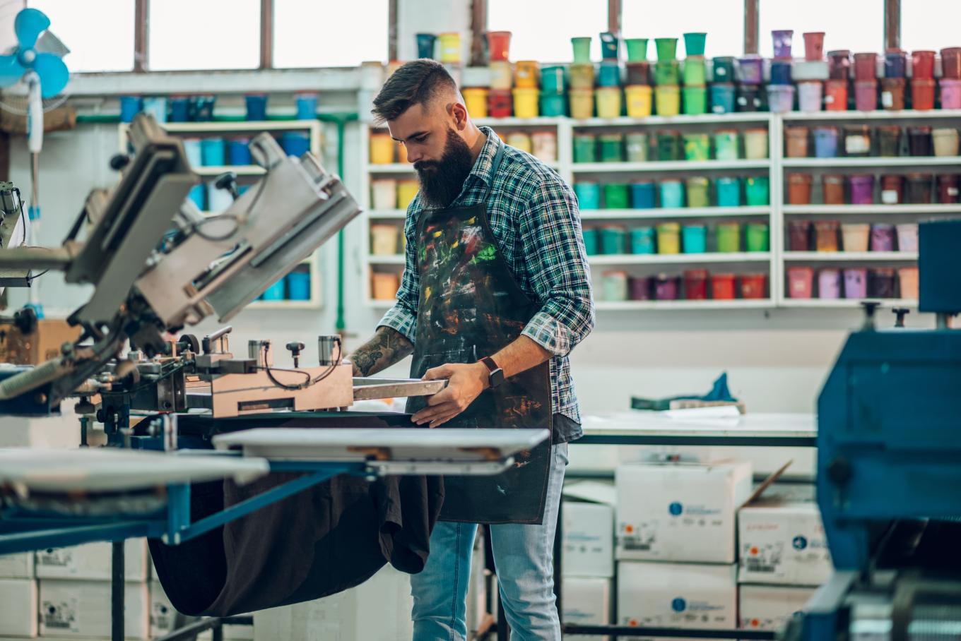 An artist surrounding by shelves of paint standing and operating a printing machine