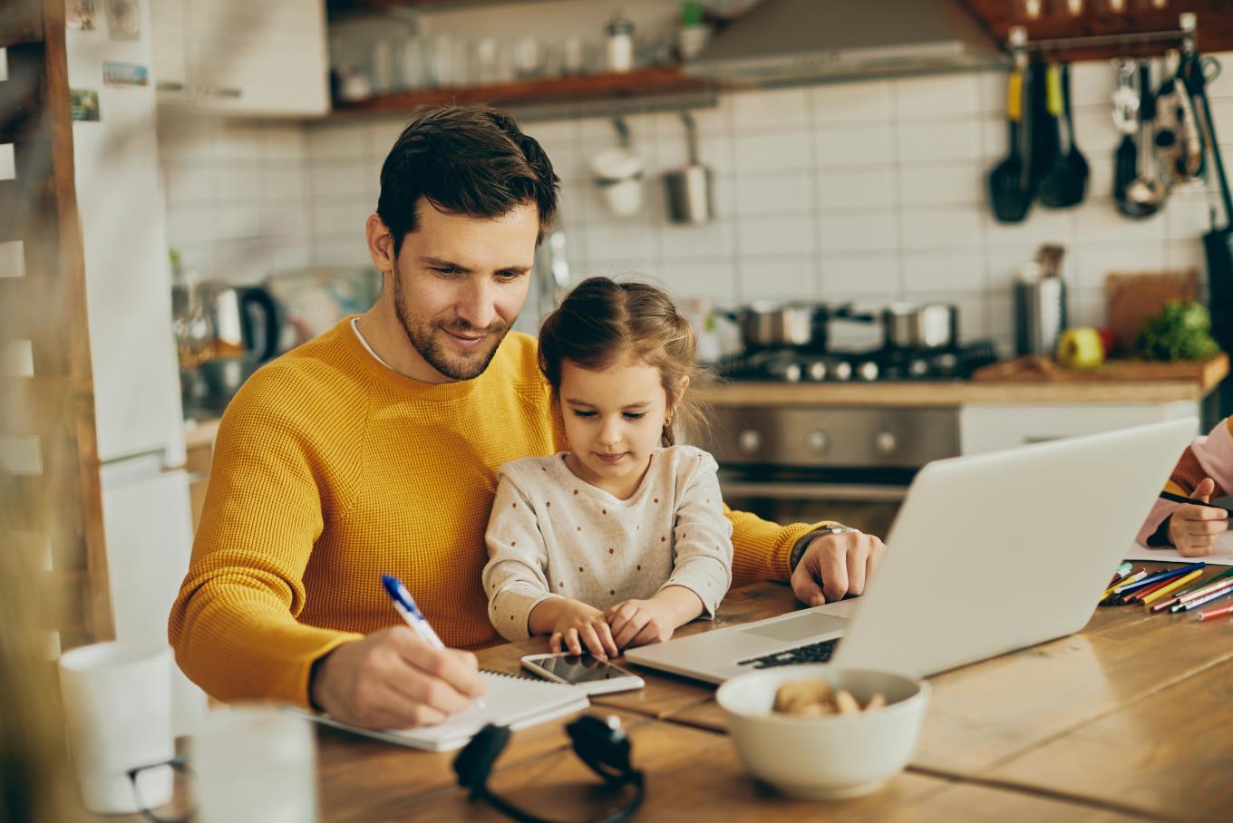 A man with his daughter sat on his lap in a kitchen sat at the dining room table with a laptop