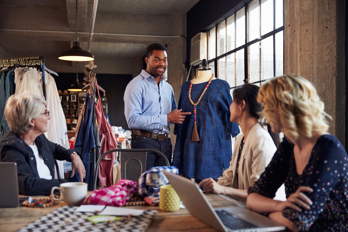 A tailor standing next to a mannequin presenting to a table of three ladies