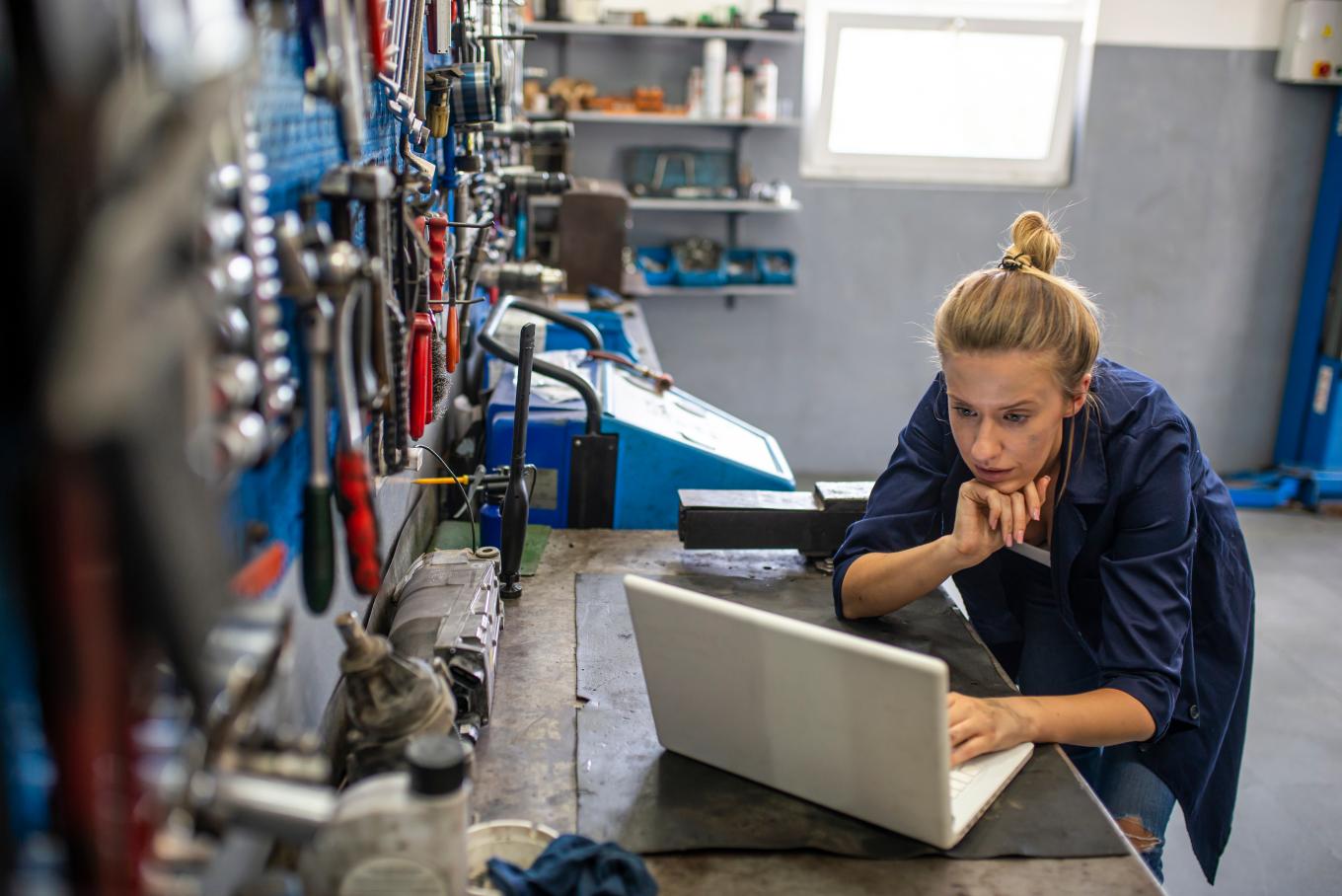 A female sat at a desk wearing overalls using a laptop in a workshop environment with walls covered in hanging tools