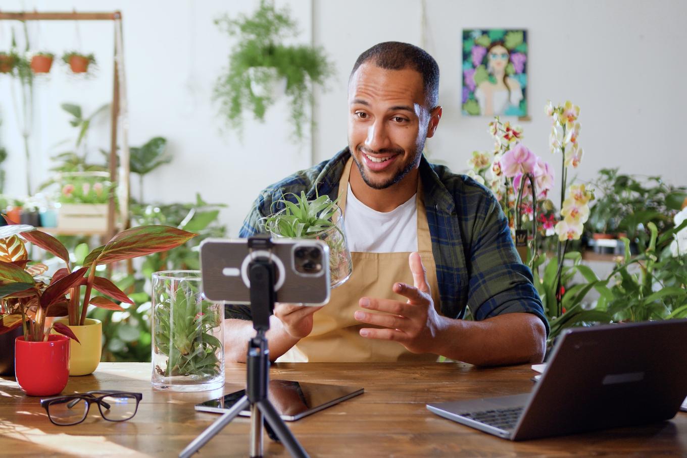 A vlogger sat at a desk filming surrounded by plants and flowers