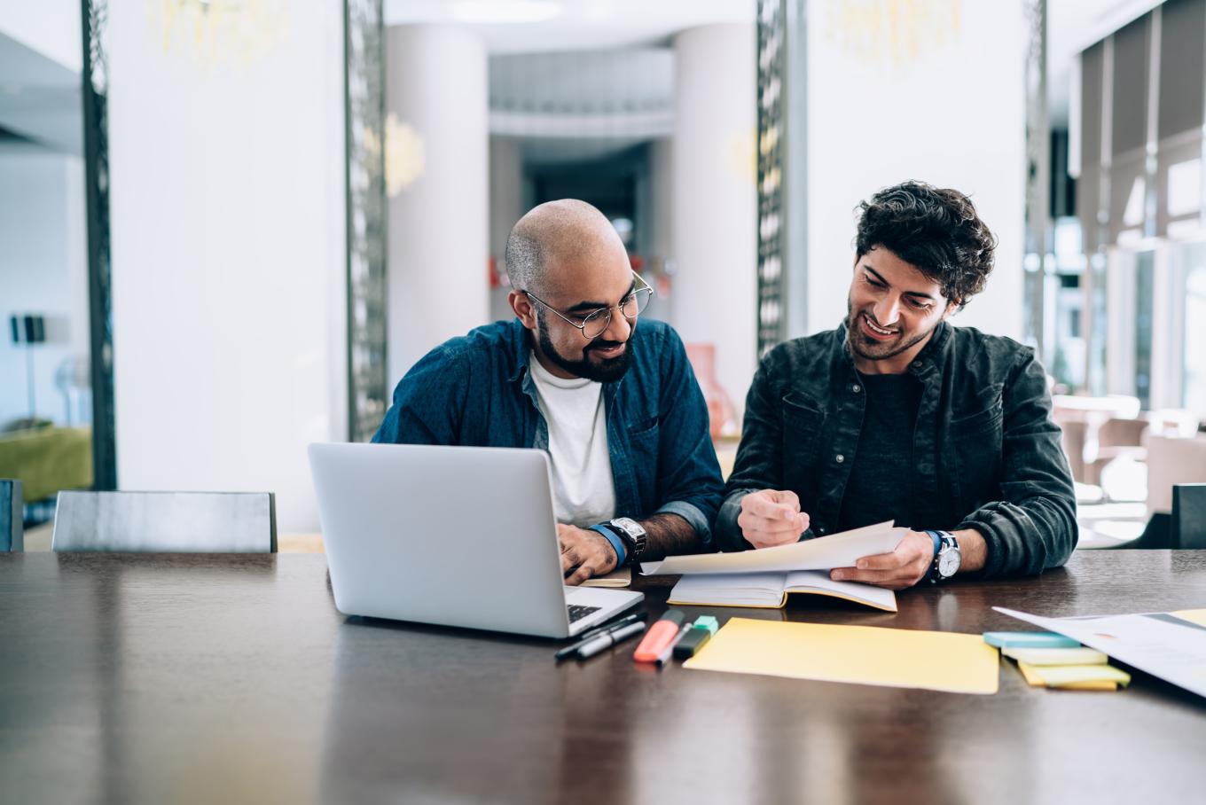 Two gentlemen sat at a table at a single laptop reviewing papers in a modern office environment
