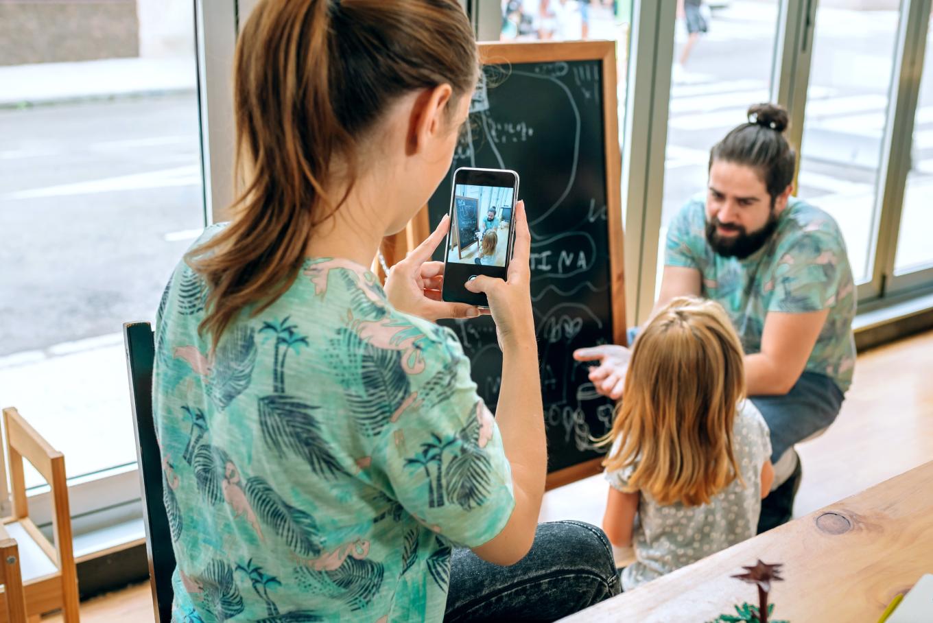 A male teacher explaining something written on a blackboard to a pupil whilst a female teacher takes a photograph